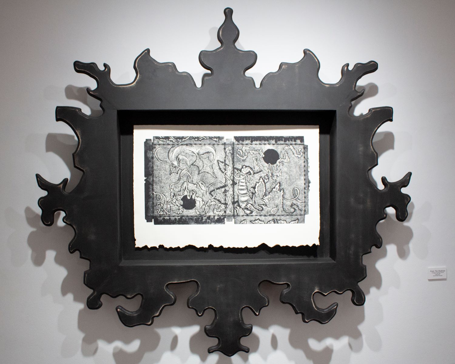 From The Shallows horizontal, greyscale Collagraph print with burnt paper edges. Large face frame with ornately shaped design elements. Rich black circles creating a diagonal within the imagery. Twisted branches/vines throughout composition. Wave splash overlapping the prehistoric figure.