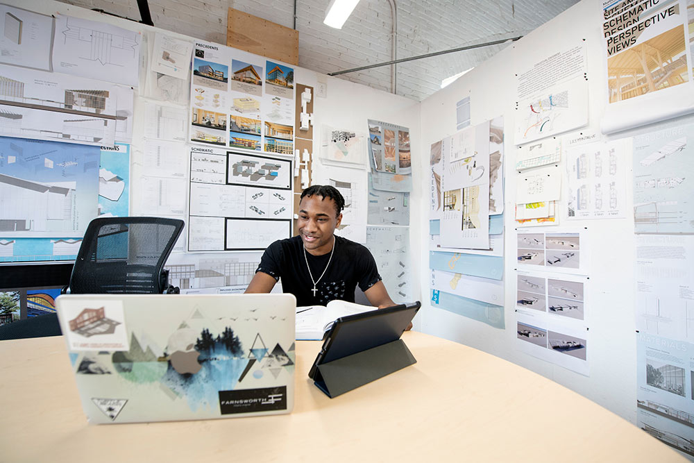an architecture student works at his computer with architecture work on walls behind him
