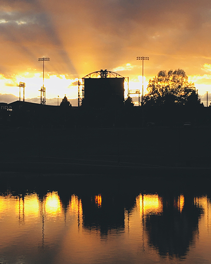 Color photograph of a yellow and gold sunset behind a sports stadium in silhouette.