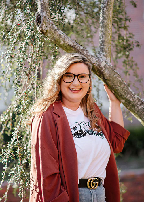 Chelsea Smith Sisson holds left arm on branch in front of brick building