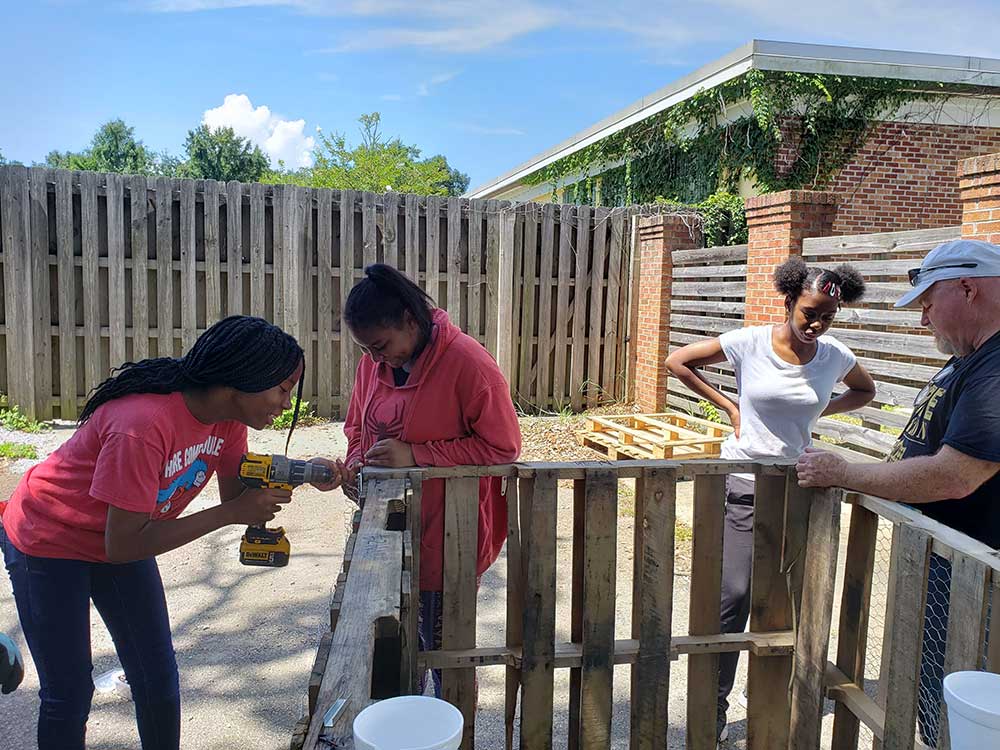Student interns (left to right) Trinity Wilson, Kiana Han and Tasia Williams work with Steve Barney to build a compost bin for the Boys and Girls Club. Shown drilling into a wood-framed box, approximately 5ft. x 5ft