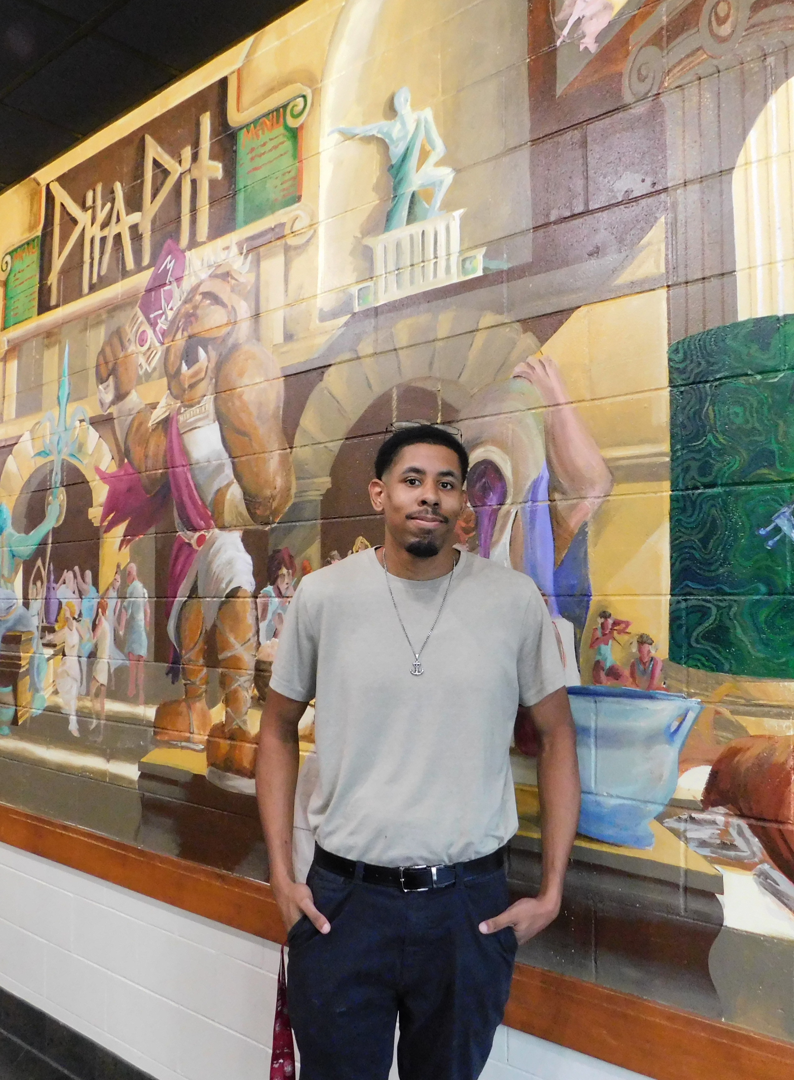 Artist Brad James Jr. stands in front of his mural, "Greek Life at Pita Pit" mural (2025)