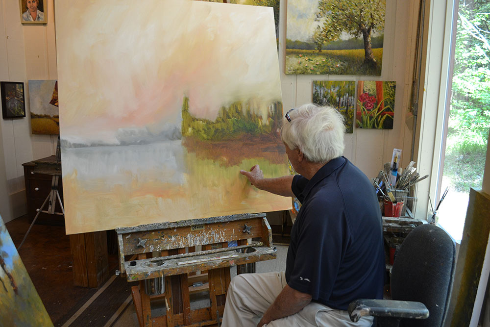 Dr. Charles Guess points to one of the two paintings in progress he worked on the day before this photo was taken.