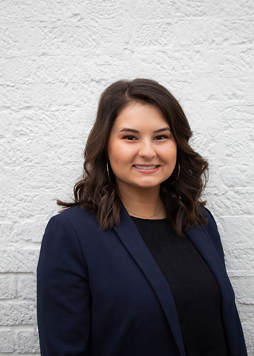 Darby Myers headshot - wearing navy suit jacket on white brick wall