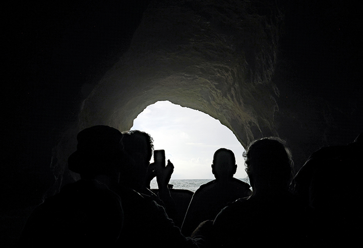 Black and white photograph of a group of people standing at the front of a cave entrance looking out at the light. One figure is lifting up a cell phone to take a picture.