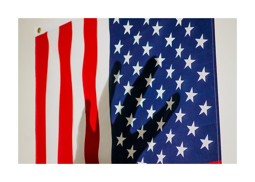 A photograph of the American flag hanging on a. wall with the shadow of a hand on top of it