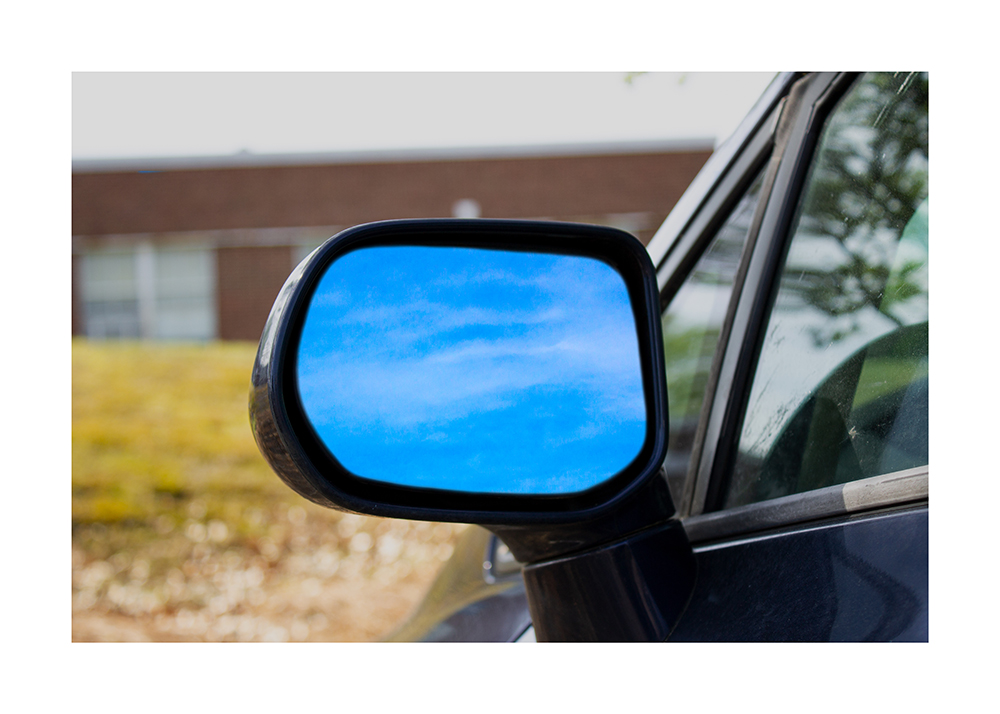 A photoshopped photograph of a car door mirror with a bright blue sky in the mirror.
