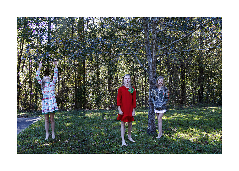 A photoshopped photograph of an image of a little girl, three versions in three outfits. The far left one is hanging from a branch, the middle one is sticking her tongue out, and the right one  is laughing.