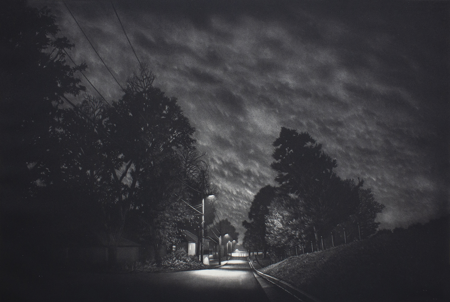 Black and white image of a dark neighborhood street lit by street lamps at night. The sky if full of clouds reflecting the light of the street lamps spotlighting the street. A row of houses line the street on the left side of the road.