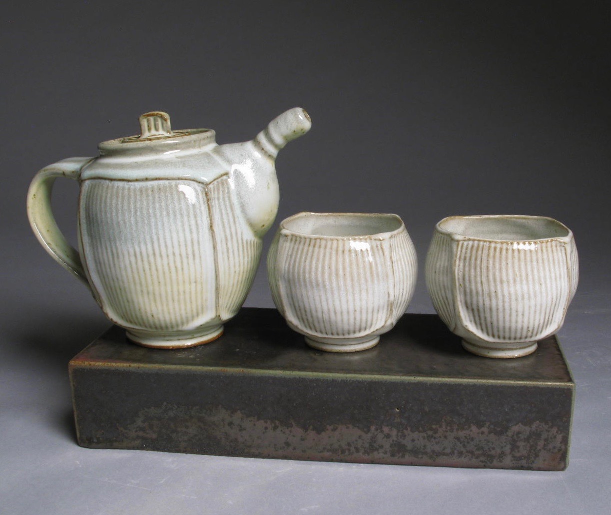 A white ceramic tea pot and two cups on a dark ceramic brick.