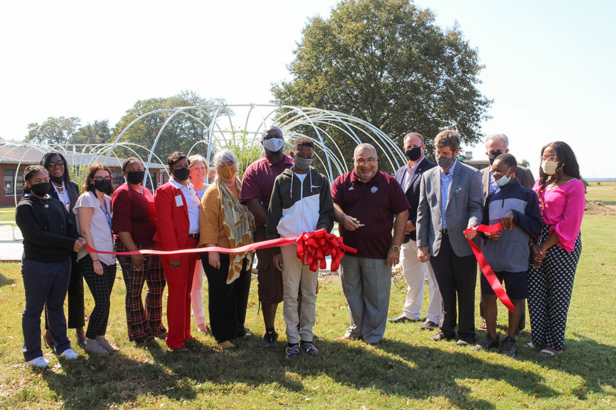 People stand in front of new community garden cutting ribbon: from left, Esperanza Colon, student; Barbara Lucas, principal; Emiline Brown, science teacher; Jamesha Keithley, science teacher; Susie Williams, district curriculum coordinator; Allison Poindexter, Delta Health Alliance; Suzanne Powney, MSU associate professor of art; Alexis Hamilton, MSU Extension agent I; Caleb Davis, student; Rev. Jesse King, district superintendent; Scott Willard, interim dean of MSU’s College of Agriculture and Life Sciences; Cory Gallo, landscape architecture professor; Sadik Artunç, professor and head of MSU’s landscape architecture department; Marquis Wafford, student; and Rickeya Brown, science teacher.