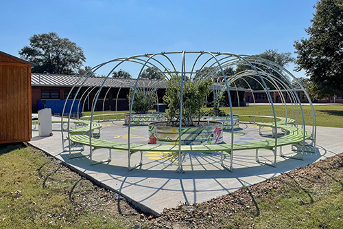 The learning garden at Leland School Park shows metal circular structure with green benches and plants on painted concrete