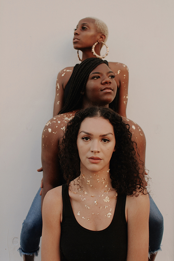 Color photograph of three female figures standing behind each other and facing the camera.