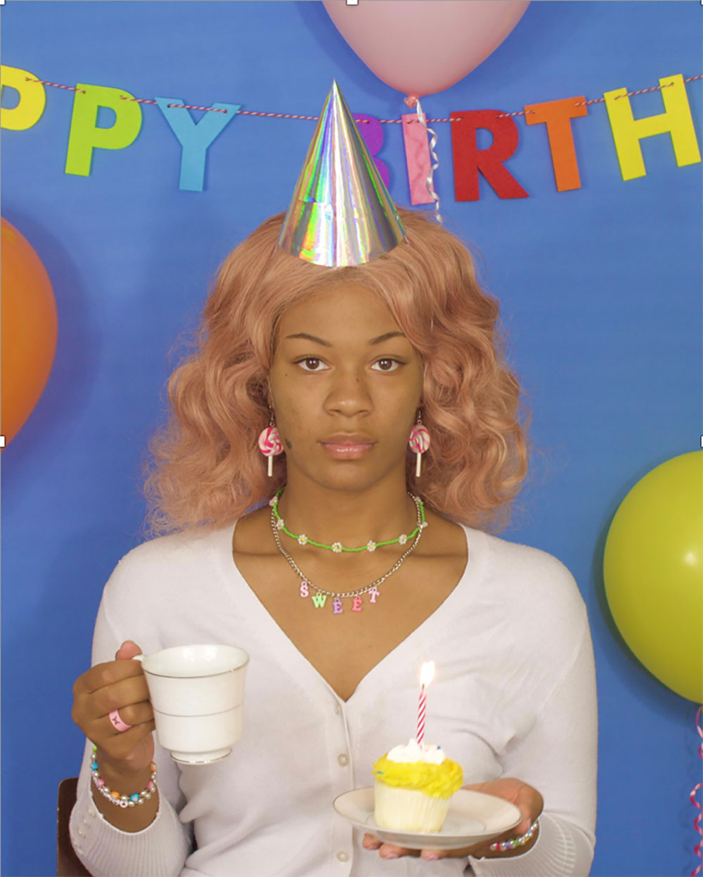 An image of a girl at a birthday party with a cupcake, tea, and a party hat - but is not smiling. 
