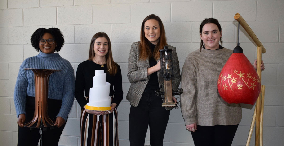 four females with their lamps: left lamp looks like a copper cylinder that angles out in a circle at top and bottom; left middle lamp looks like a white layered wedding cake; right middle lamp looks similar to an old rotary phone; right lamp looks like a red buoy with yellow flowers on it hanging from a wooden base 