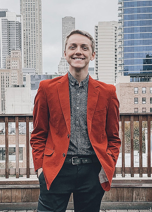 Houston McMahon stands with city scape in background - wearing red(ish) suit jacket