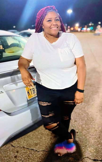 Photograph of a young woman leaning on the back of a car parked in a parking lot at night.