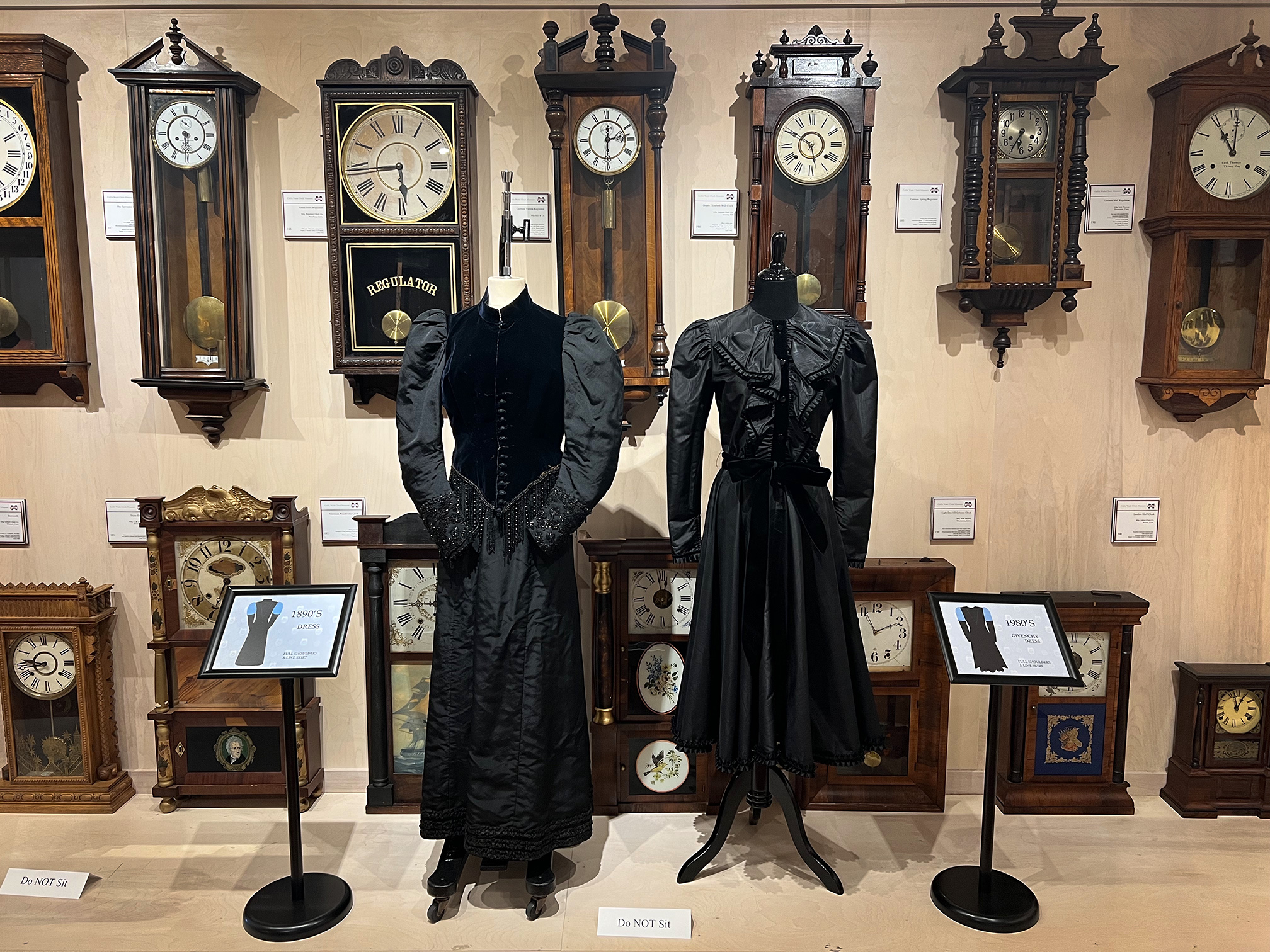 Two black dresses on display in front of a row of old clocks.