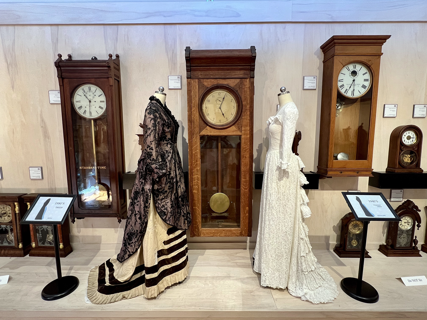 Two historic dresses on dress forms in front of historic wooden clocks.
