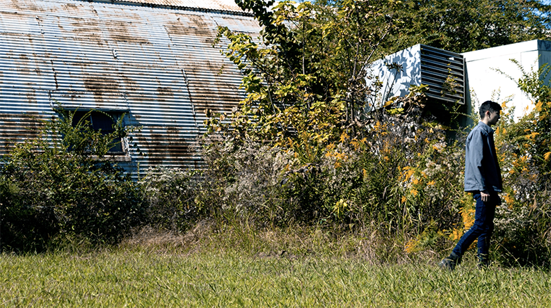Still photography from a short film. Color image of a figure walking in front of an old rusted metal building.
