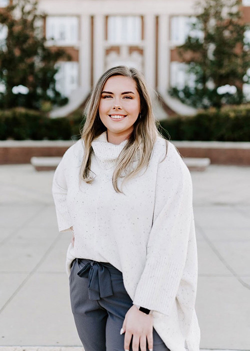 Katie Vandiver in white sweater poses in front of MSU campus building