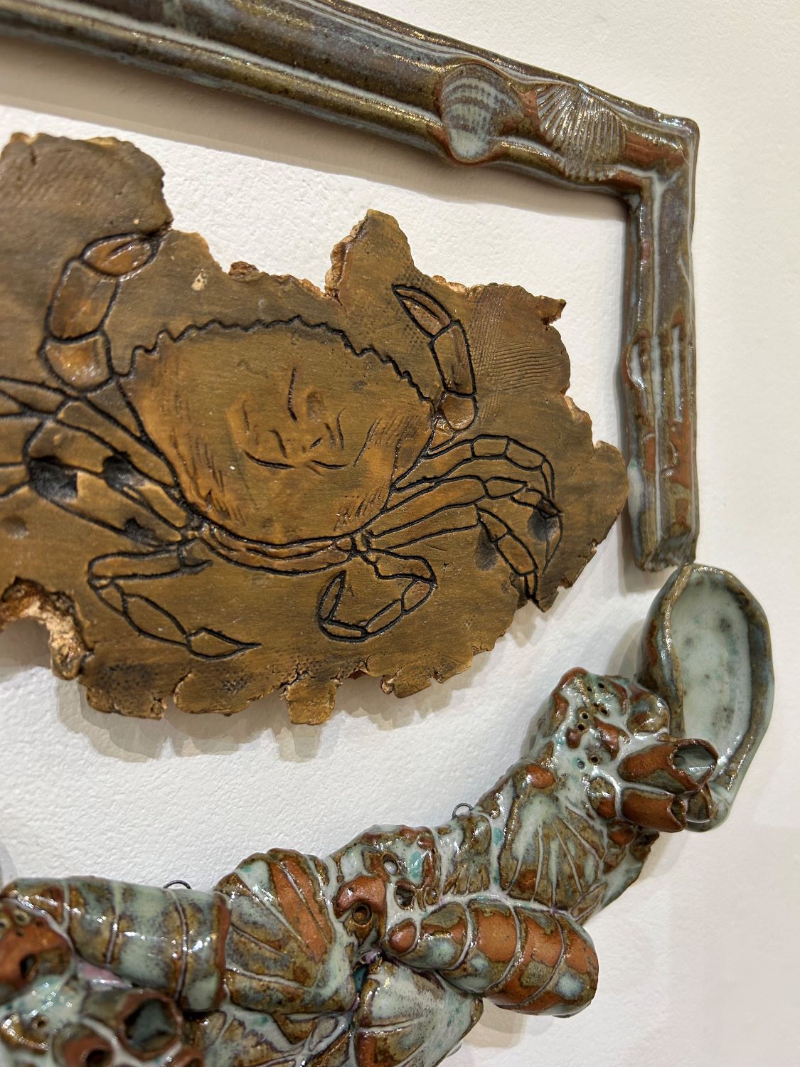 Detail of Carcinus maenas 1, depicting the European Green Crab on a stoneware slab surrounded by frame with seashells made with stoneware clay, glazed in Seafoam Green. 