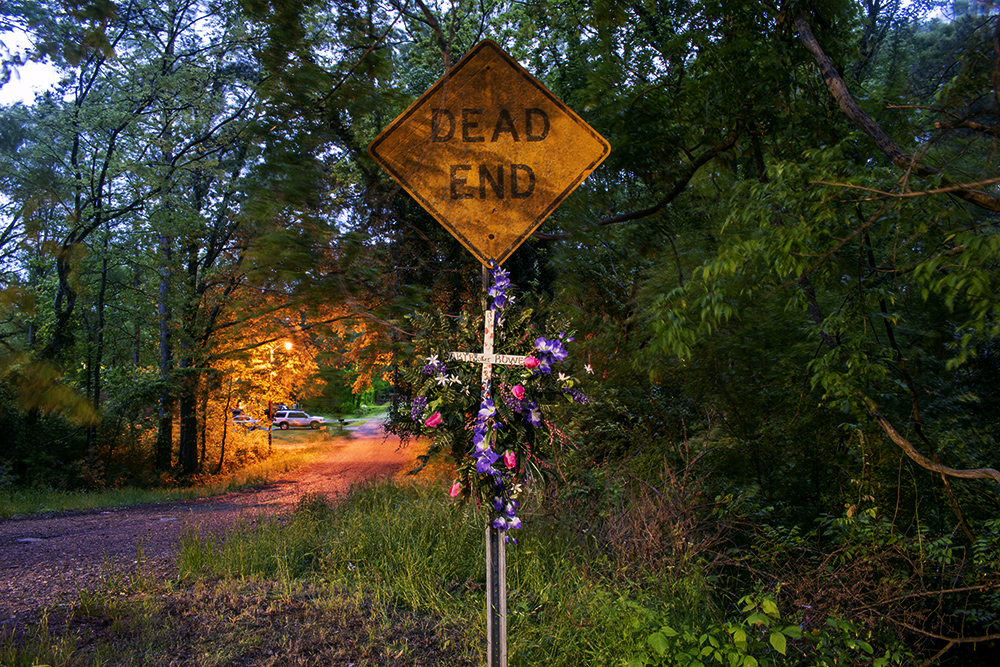 A photograph of a dead end sign with a cross attached to it surrounded by flowers