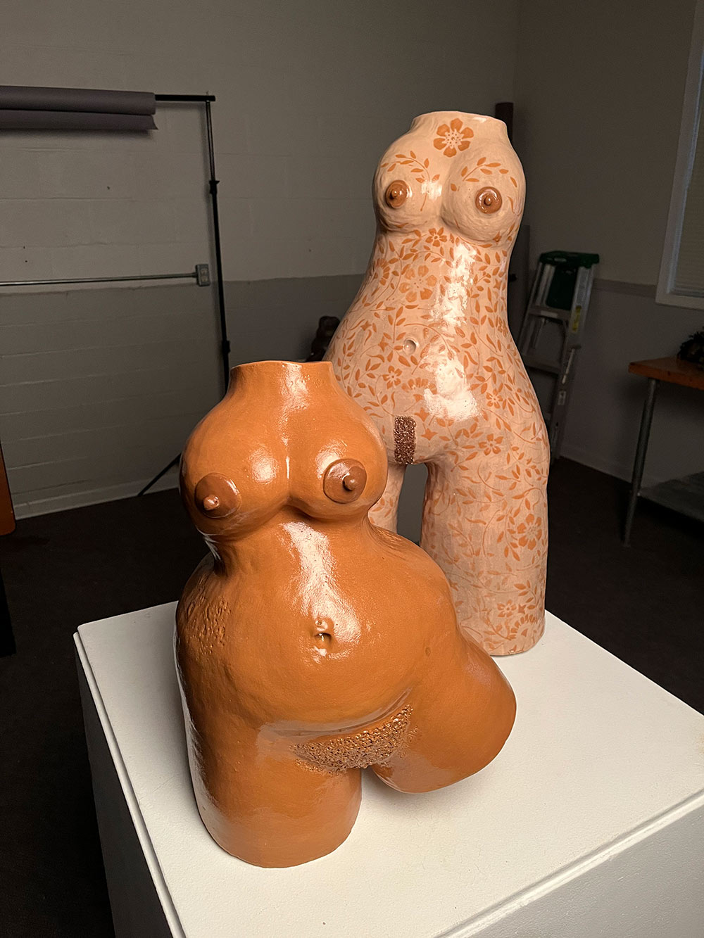 A small fleshy brown sculpture with nipple and belly rings and A tall fair skinned sculpture with brown floral tattoos covering the entire piece