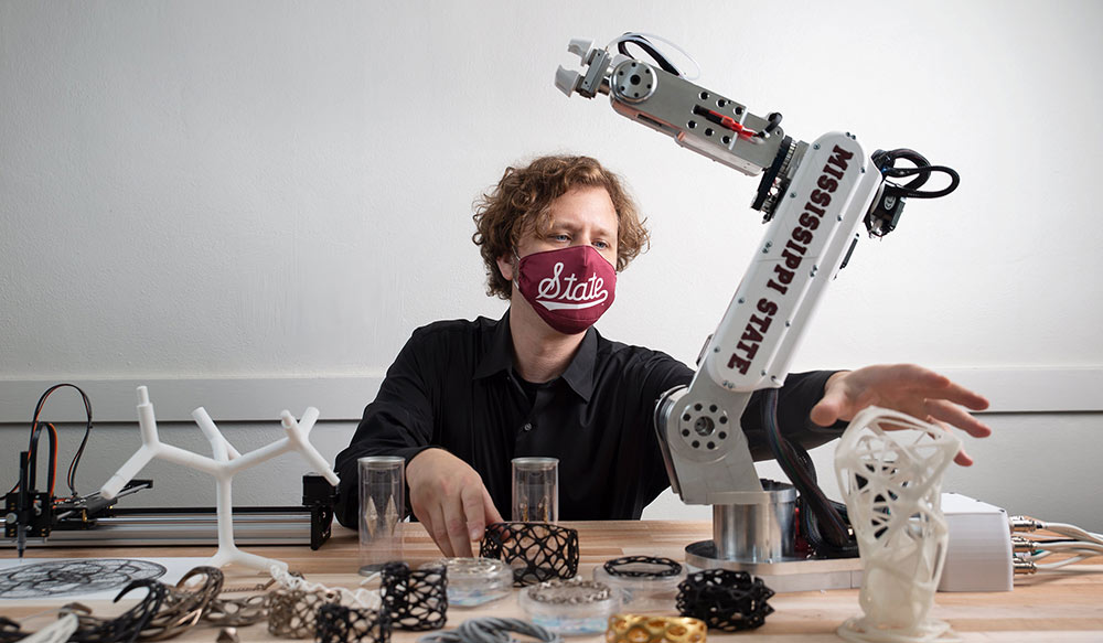 Duane McLemore wearing a MSU mask works on his desk with a robot and 3d printed material