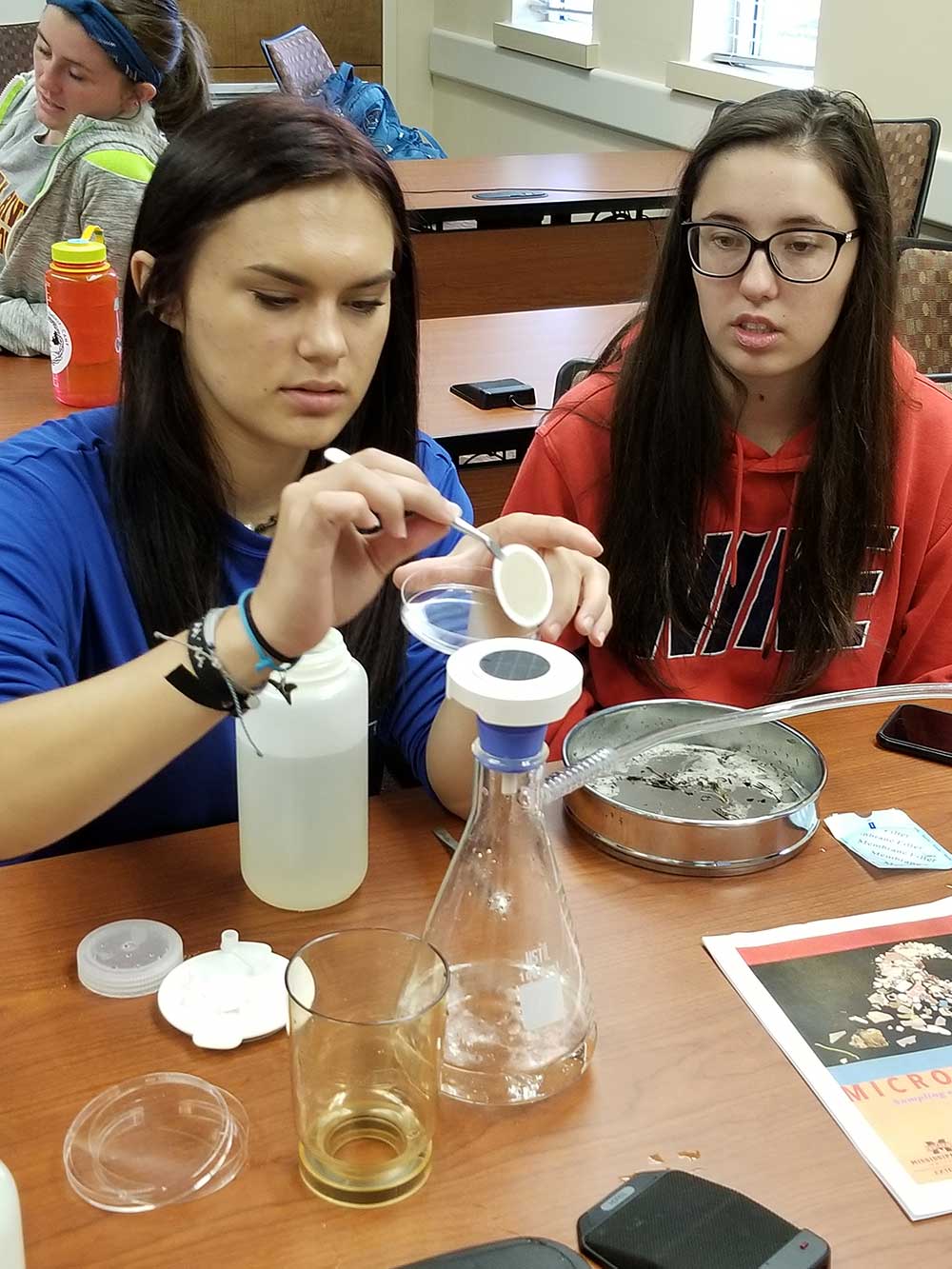 nterns analyzing microplastics as part of Student Master Naturalist Program. Two students work with science materials (beaker, etc. at a table)
