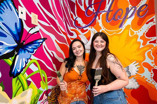 MSU senior art majors Gabby Castleman, left, and Sara Beth Messemore pictured with their mural which incorporates symbols of hope and recovery. (Photo by Emily Grace McCall)