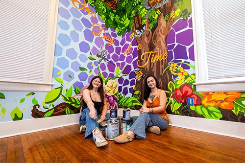 MSU senior art majors Sara Beth Messemore of Madison, left, and Gabrielle “Gabby” Castleman of Brandon, sit next to the mural they painted this summer in MSU’s Bulldogs in Recovery lounge, which is part of the Office for Student Well-Being. (Photo by Emily Grace McCall)