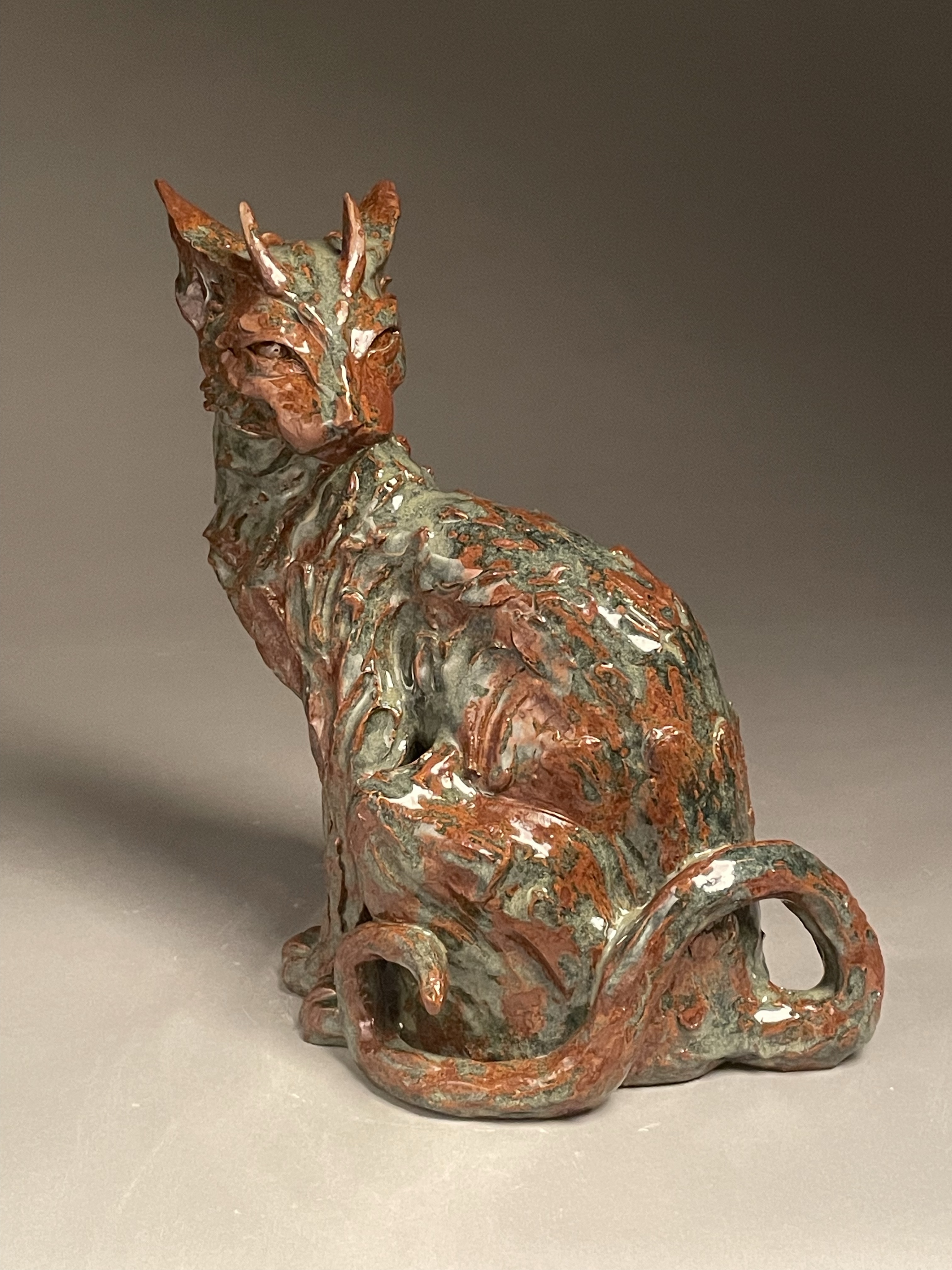 Ceramic cat sculpture with horns sitting and looking behind itself with a blue green glaze