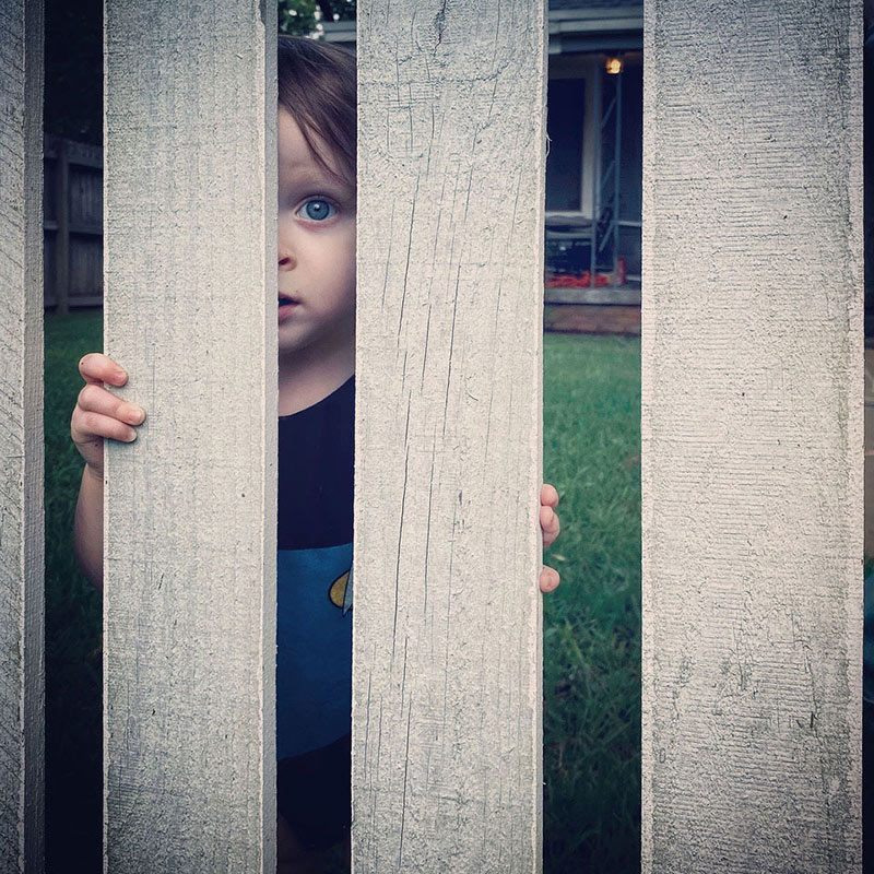 "Six Feet From Grandma" photograph by Marita Gootee - shows young child peeking through a gray fence