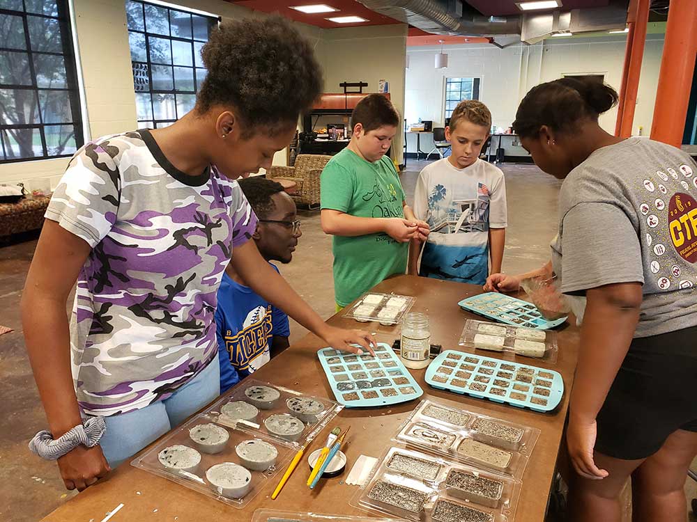 Students at the Hancock County Unit of the Boys and Girls Clubs learning how to make organic soap. People are gathered around a table with soap in molds of various shapes and sizes