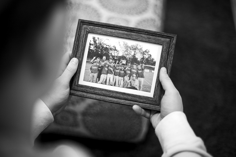 A black and white photographed image of a child holding a framed picture of a youth baseball team.