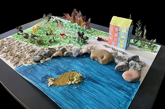 Sculpture on a poster board with water, rocks, and a building.