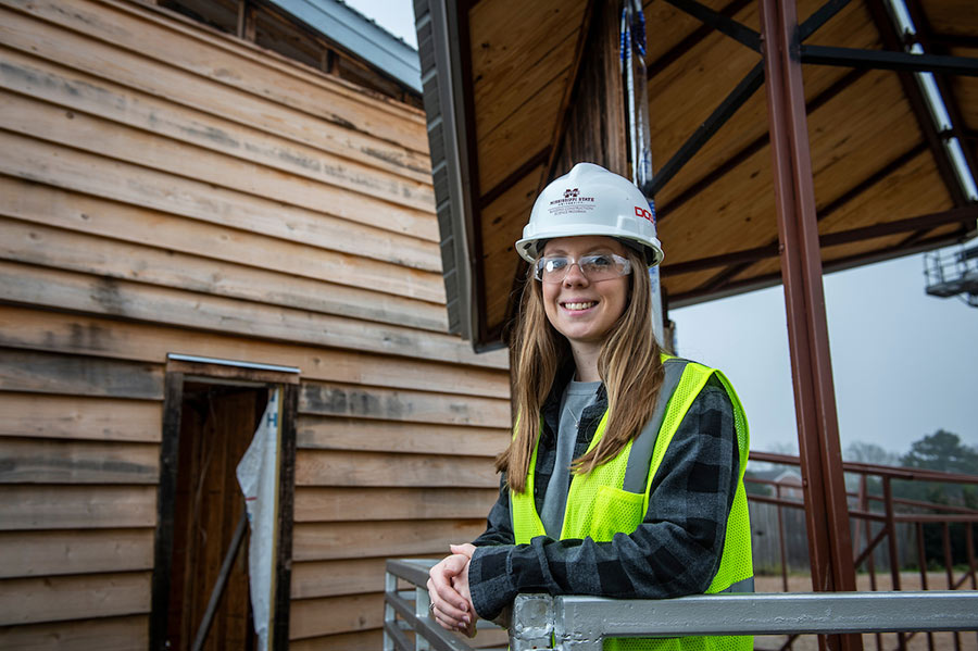 Jalyn Wallin stands wearing hard hat and vest in front of modular building project