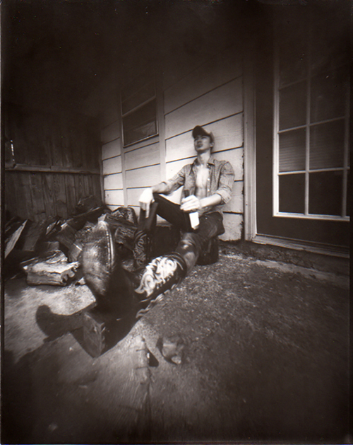 Black and white photograph of a figure wearing cowboy boots and sitting on the ground, leaning against the outside of a building. The image is blurred.