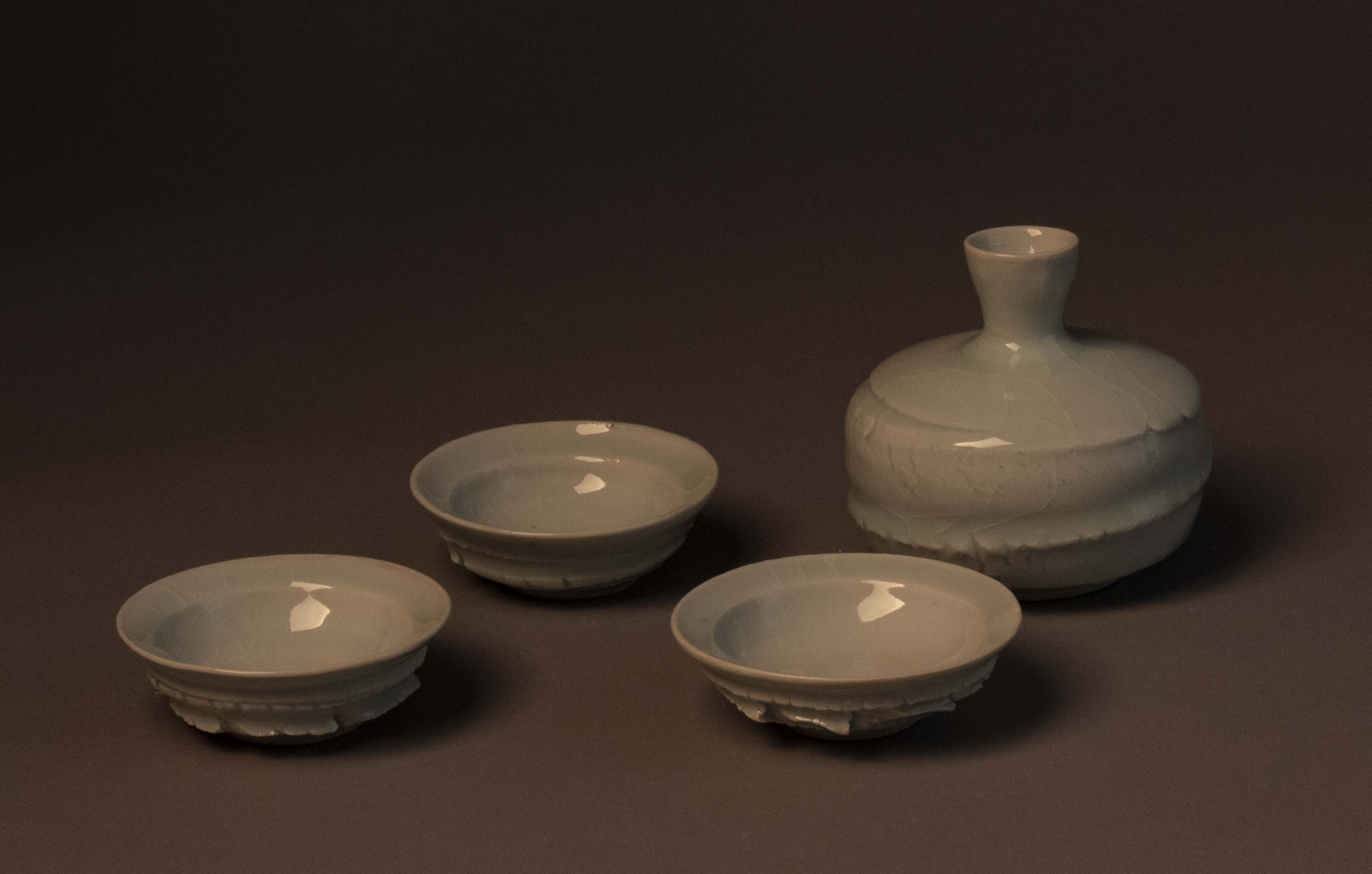 Three small saucer-shaped cups, and a vase-like bottle. The interior surfaces of the shallow cups (guinomi) are smooth while the exterior bottom portions of the cups have a texture that might almost feel jagged like chipped stone. The bottle (tokkuri) has a squat almost barrel-like shape with a neck that is narrow at the base and flares out at the lip. While the neck and shoulder of the bottle are smooth, the body portion of the vessel has some the same stretched clay textures as the exterior of the cups. The glaze is glassy and relatively smooth, though thin cracks can be seen in the glaze (crazing).