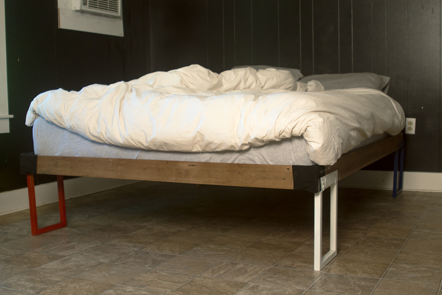 side angle view of bed frame, steel rectangle legs, with leather detail on corners. ruffled comforter on top.