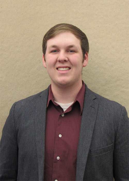 Black Farrar wearing maroon dress shirt and gray jacket stands against light tan wall inside