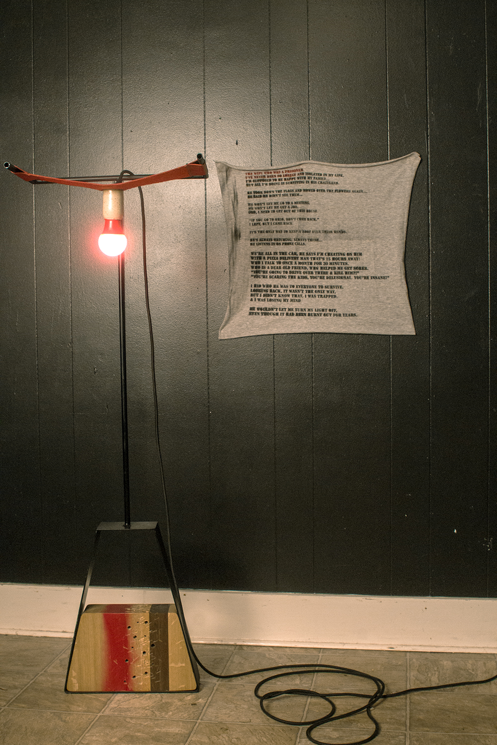 metal lamp with trapezoid base with wood in center, light housing is ceramic in leather sling. with small tapestry behind with poem on it. 