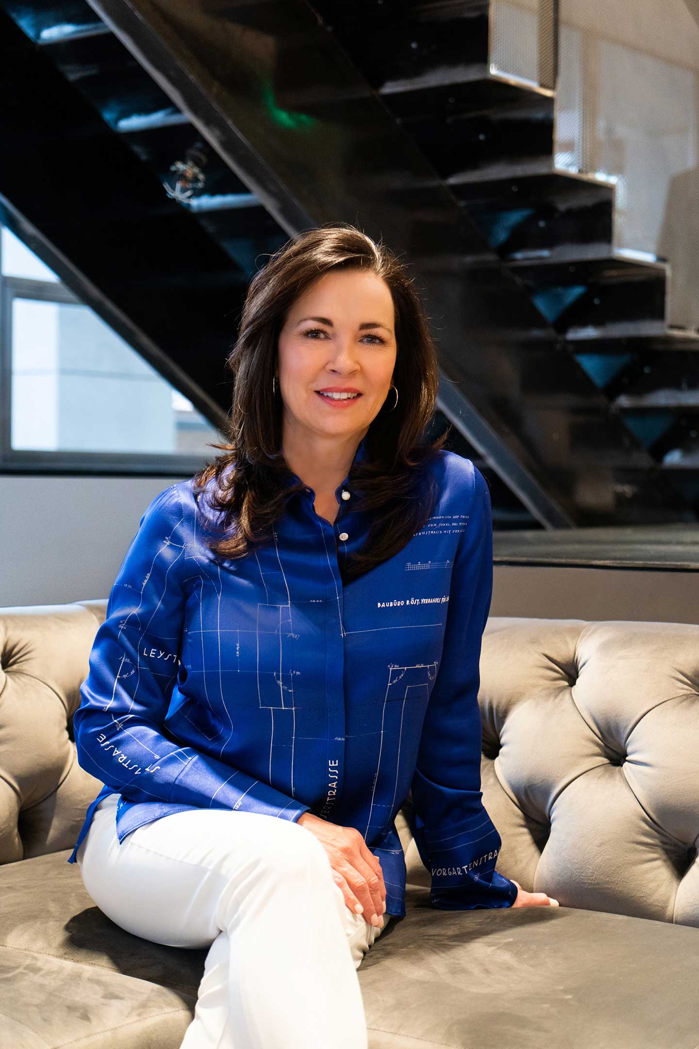 MSU interior design alumna Cindy Simpson, IIDA Fellow - Cindy sits on a gray suede couch. She is wearing a blueprint blouse and white pants.