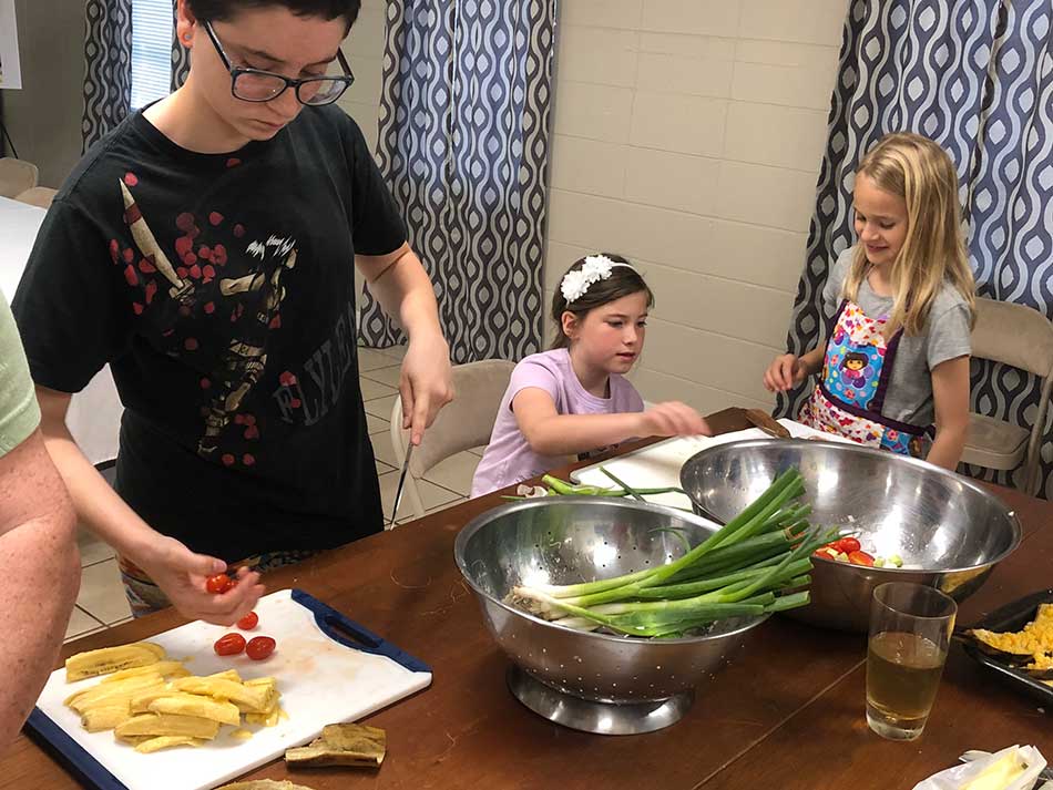 children prepare food around a table - green onions, plantains, and cherry tomatoes are shown