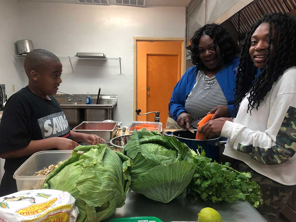 A boy, left, and two women gathered around a table with vegetables - possibly making a salad. Carrots, lettuce or cabbage, and cilantro are on the table.