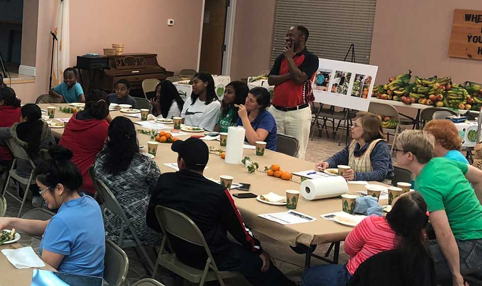 shot of a room of people sitting at large tables - bananas or plantains and possibly apples are piled on a table in the back of the room. Oranges or tangerines are on the table as a center piece.